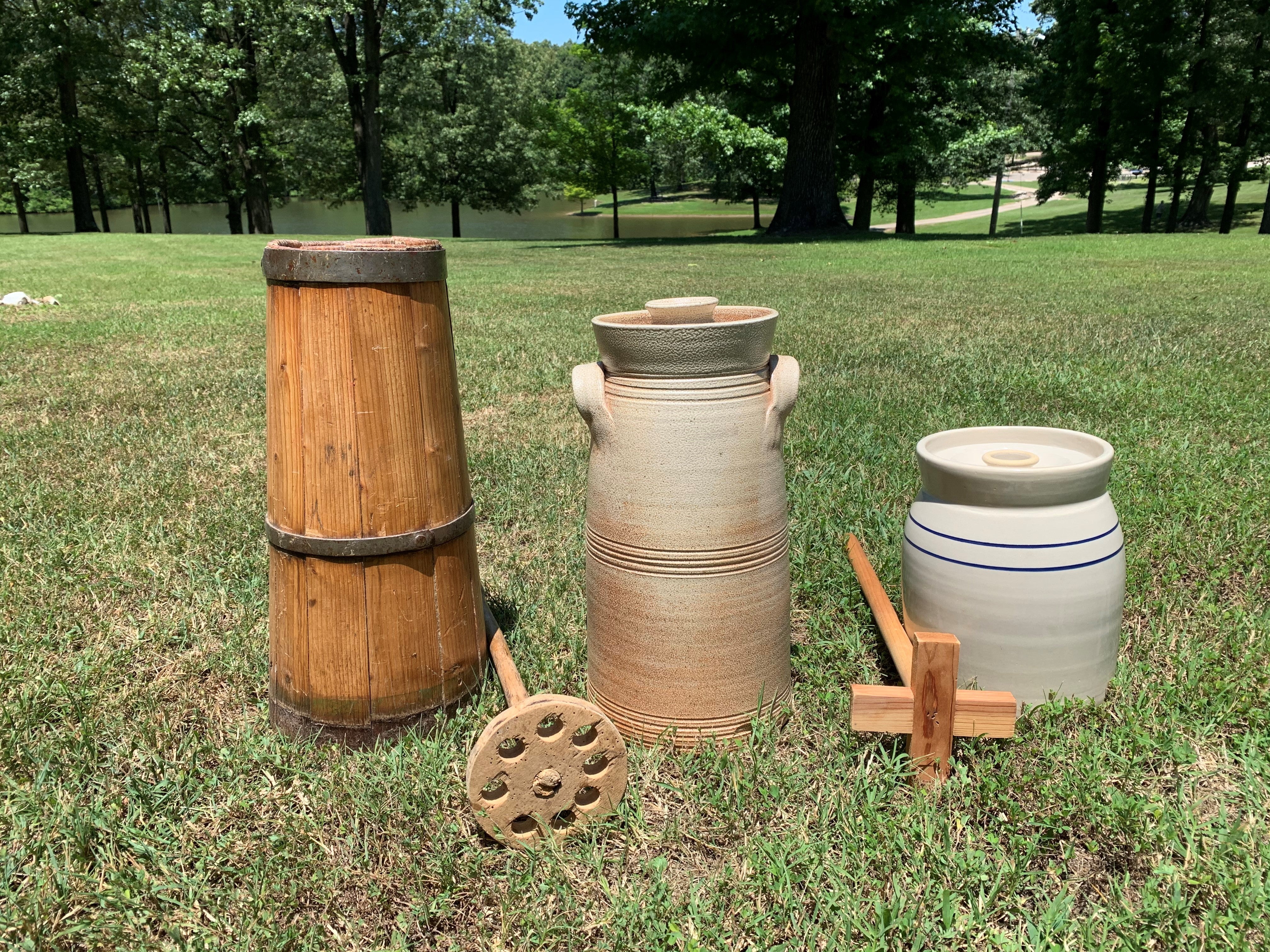 Three butter churns are lined up outside from tallest to shortest. The first churn is wooden and the other two are clay. There are two dashers (wooden stick used to churn the butter) in between the three churns. One dasher has a cross pattern and the other is a circle with holes in it.)